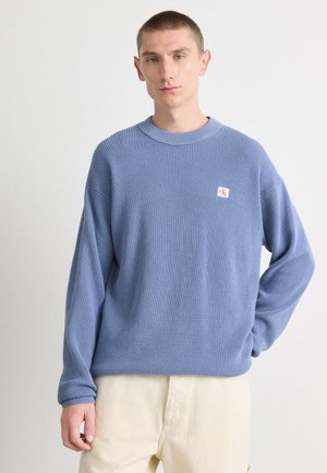 Lichtblauwe wafelgebreide trui met een ronde halslijn en verlaagde schouders. Bevat een klein logo-patch op de borst. G gedragen met beige broeken.