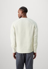 Chunky gestrickter cremefarbener Pullover mit geripptem Textur, rundem Ausschnitt und überschnittenen Schultern. Kombiniert mit grauen Jeans.