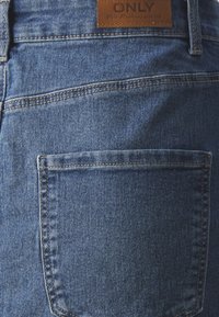 Középkék mosású farmernadrág, hátulsó zsebbel, látható varrási részletekkel és "ONLY" márkájú barna bőrcímkével.