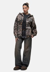 Giacca con motivo camouflage dotata di cappuccio in tonalità marrone e grigio, abbinata a pantaloni in denim scuro e scarpe marroni, con una vestibilità comoda.