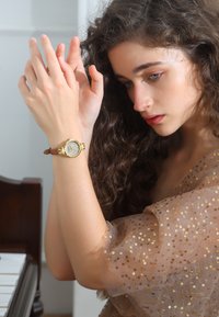 Montre dorée avec un cadran rond et un bracelet en cuir marron. Le bracelet est fixé sur un poignet orné d'une manche transparente embellie de dorures.