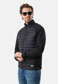 Man wearing black quilted jacket and sunglasses, standing with hands clasped, looking slightly to the side.