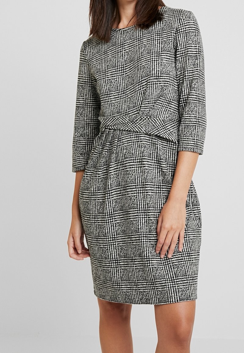 Femme portant une robe à carreaux noire et blanche longueur genou avec des manches trois-quarts et une taille cintrée.