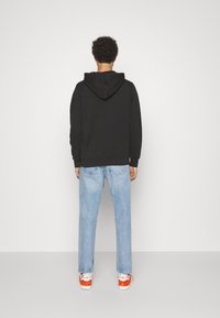 Sudadera con capucha negra, de corte holgado, combinada con jeans de color azul claro. Lleva zapatillas naranjas con acentos blancos y un logo rojo; puños visibles.