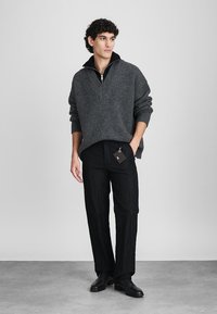 Grauer Strickpullover mit einem halben Reißverschluss-Kragen, kombiniert mit strukturierten schwarzen Hosen. Das Outfit hat ein Preisschild angebracht. Schwarze Schuhe vervollständigen den Look.