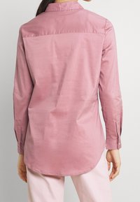 Light pink long-sleeve shirt with a collar and buttoned cuffs. Smooth texture with a slightly curved hem and yoke detail at the back.