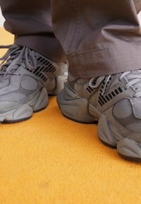 Šedé športové topánky s ktorými sú kombinované sieťové a semišové textúry, vybavené robustnou podrážkou a čiernymi pruhmi. Nosí sa s šedými nohavicami na oranžovom povrchu.