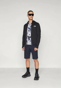 Άντρας που φοράει μαύρο φούτερ με φερμουάρ, μπλούζα καμουφλάζ σε μπλε και άσπρο, ναυτικές βερμούδες, μαύρες κάλτσες, μαύρα αθλητικά παπούτσια και μεγάλα μαύρα γυαλιά ηλίου, στέκεται μπροστά από έναν λείο φόντο.