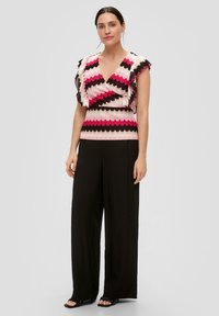 Multikleurige top met zigzagpatroon in roze, roomkleur en zwart, met gerimpelde mouwen, gecombineerd met zwarte broek met wijde pijpen.