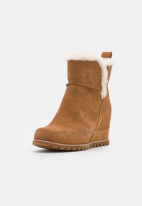 Botte en suède beige avec semelle compensée, doublée de fausse fourrure blanche, à bout arrondi et semelle en caoutchouc texturé. Équipée d'une languette à l'arrière pour un enfilage facile.