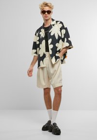 Chemise à manches courtes florale en bleu marine et crème, associée à un short crème clair. Des chaussures noires et des lunettes de soleil complètent la tenue.