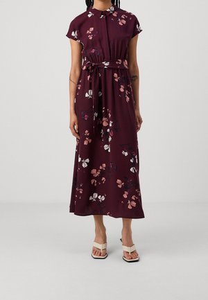 Kobieta ubrana w bezrękawnikową marszczoną sukienkę midi w kolorze bordo z kwiatowym wzorem, z paskiem wiązanym w talii i beżowymi sandałami na obcasie, stojąca na jednolitym tle.