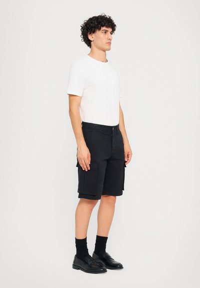 Schwarze Cargo-Shorts mit mehreren Taschen, aus Baumwollstoff, kombiniert mit einem schlichten weißen T-Shirt und schwarzen Slippern. Das Modell steht seitlich.