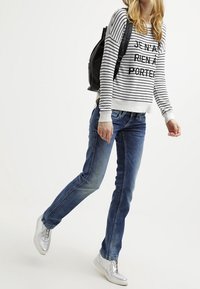 Suéter a rayas blanco y negro con texto, jeans de mezclilla azul, y zapatillas altas plateadas. Mochila llevada en un hombro.