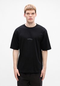 Joven con cabello corto que lleva una camiseta negra de gran tamaño con el logo "HUGO" en el pecho, de pie contra un fondo blanco liso.