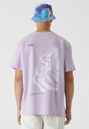 Lilac cotton t-shirt featuring a fluid white abstract graphic on the back, paired with a multicolored tie-dye bucket hat.