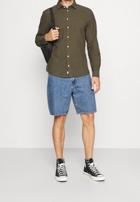 Camisa de botones verde oliva con cuello, pantalones cortos de mezclilla azules y zapatillas altas negras, sosteniendo un pequeño bolso negro de hombro.