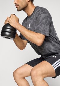 Haltère noir lesté tenue par une personne portant un t-shirt gris à motifs avec un col rond foncé, et un short noir avec des bandes blanches.