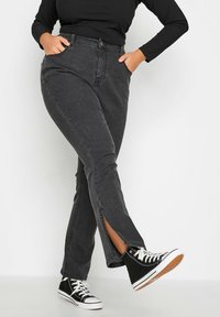 Czarne, wysokie dżinsy z dopasowanym krojem, z przednimi kieszeniami i bocznymi rozcięciami u dołu, noszone z czarnymi trampkami typu high-top.