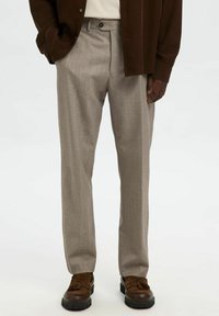 Man som bär ljus beige skräddarsydda byxor, mörkbruna loafers med fransar, en mörkbrun skjorta och en stoppad beige topp.