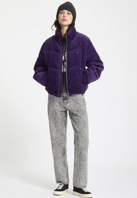 Paarse corduroy puffer jack met hoge kraag, voorzien van een ruitvormig quiltpatroon. Gedragen over een grijze shirt en gecombineerd met grijze jeans.