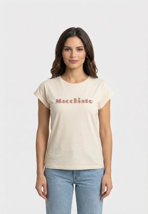 Donna con capelli lunghi e scuri che indossa una maglietta beige con maniche arrotolate con la scritta "Macchiato" e jeans azzurri chiari su uno sfondo bianco semplice.
