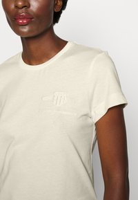 Camiseta de color beige claro hecha de una tela suave. Presenta un cuello redondo, mangas cortas y detalles en relieve del logo en el pecho.