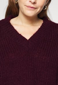 Burgundy stickad tröja med V-ringning, som har en ribbad textur och mjukt tyg, med intrikata stygn detaljer.