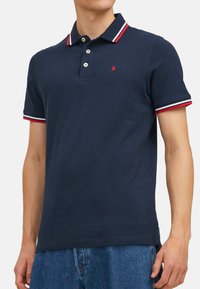 Marineblauwe polo met witte en rode gestreepte kraag en mouwafwerking. Voorzien van een knoopsluiting met drie knopen en een klein logo op de borst. Zachte stof.