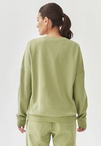 Felpa verde con una vestibilità rilassata, colletto girocollo, spalle scese e polsini a coste. Texture morbida con una superficie liscia.