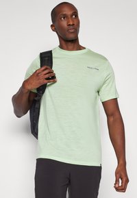 T-shirt verde-claro de gola redonda, feita de um tecido texturizado, com um design simples e um pequeno logotipo na frente. Usada com uma mochila preta.