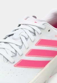 Λευκό αθλητικό παπούτσι με ροζ λεπτομέρειες, διαθέτει τρεις εμβληματικές ρίγες, υφασμάτινο επάνω μέρος με υφή και επίπεδα κορδόνια. Στερεή σόλα από καουτσούκ.