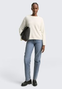 Weißer, flauschiger Pullover mit rundem Ausschnitt, kombiniert mit blauen Jeans und schwarzen Plateauschuhen; hält eine schwarze Handtasche.