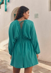 Vêtement teal à manches longues avec une taille smockée et un dos ouvert maintenu par un lien. Tissu doux et texturé ; design aéré et ample.