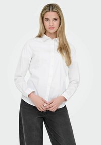 Jeune femme aux longs cheveux blonds portant une chemise blanche boutonnée et un jean noir, debout devant un fond clair uni.