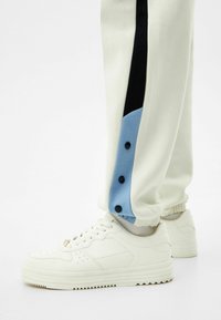 Bílé tenisky nošené s krémovými kalhotami, které mají modré a černé boční panely a tři černé knoflíky blízko kotníku.