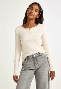 Beige långärmad topp med en vågig halslinning och tre knappar vid bröstet, kombinerad med högmidjade grå jeans. Mjuk textur.