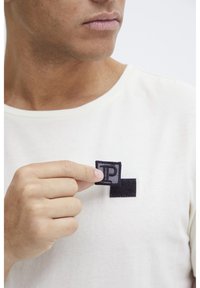 Beige t-shirt med et tekstureret sort og gråt mærke, der viser bogstaverne "P" og "T." Mærket holdes med den ene hånd nær brystområdet.