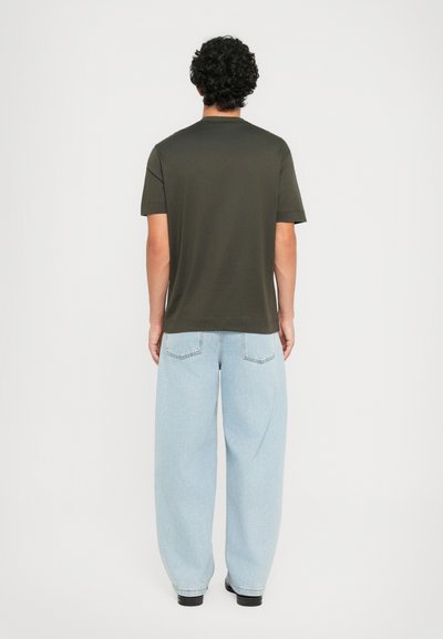 Göndör hajú férfi háttal áll, sötétzöld, rövid ujjú pólót, világoskék, bő szárú farmert és fekete cipőt visel egyszínű háttér előtt.