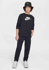 Tinédzser fekete Nike kalapban, fekete hosszú ujjú Nike pólóban, fekete nadrágban és fehér sportcipőben áll, egyik kezével a zsebében.