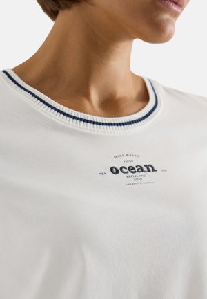 Gros plan d’une personne portant une chemise blanche avec un col rayé bleu marine et le texte « ocean » ainsi que des mots associés sur la poitrine.