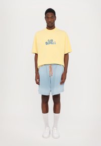 Geel t-shirt met korte mouwen met blauwe grafische tekst, gecombineerd met lichtblauwe shorts met een gemusterde trekkoord, witte sneakers en kniekousen.