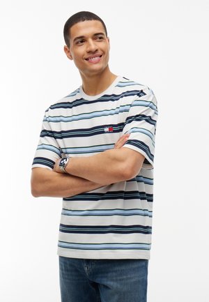 Mężczyzna uśmiecha się, z założonymi ramionami, ubrany w białą pasiastą koszulkę Tommy Jeans i niebieskie jeansy na jednolitym tle.