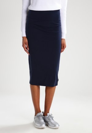 Personne portant une jupe crayon bleu marine jusqu'aux genoux, un haut blanc côtelé à manches longues, et des baskets de sport argentées, debout sur un fond uni.