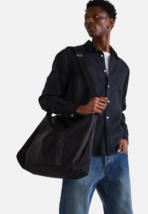 Sac en toile noir au toucher doux, avec des accents en cuir noir contrastants et un design spacieux, tenu par une personne.