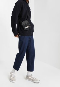 Sacoche noire zippée à logo blanc, portée sur un sweat à capuche sombre, associée à un pantalon en denim et des baskets grises. Détails en tissu texturé visibles.
