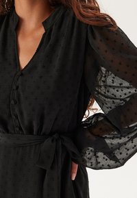 Svart klänning med V-hals, knappdetaljer och textilstruktur. Genomskinliga puffärmar med prickigt mönster, knuten midjeaccent.