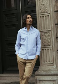 Gestreept blauw-wit overhemd met knopen, lange mouwen, losse pasvorm. Crèmekleurige broek, staand tegen een sierlijke stenen muur.