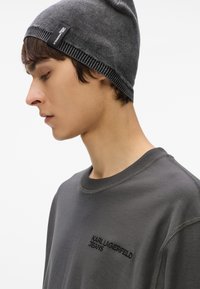 Pelēka adīta cepure ar melnu zīmola birku; pelēks džemperis ar "KARL LAGERFELD JEANS" izšuvumu; ar apaļu kaklu un teksturētu audumu.