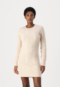 Strikket kjole i beige med teksturerte kabelmønstre, lange ermer, rund hals og ribbestrikkede mansjetter og nederkant. Fitted silhuett.